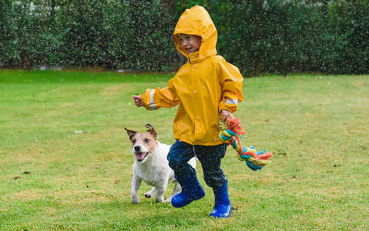 Kind springt in Regenkleidung mit Hund über eine Wiese