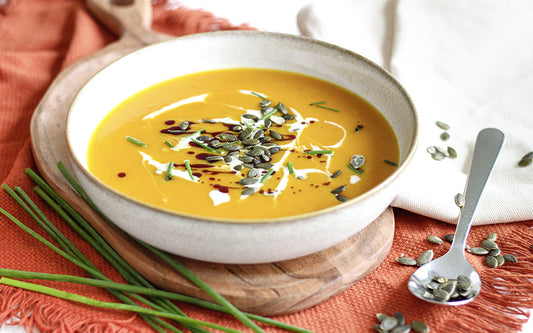 Gelbe Kürbissuppe in einer Porzellanschale. Darunter liegt ein Holzbrett und ein rotes Leinentuch. Drumherum liegen Kürbiskerne 