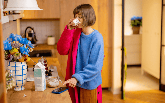 Person, mittellange braune Haare, bereitet parallel Frühstück zu, trinkt Kaffee, tippt ins Handy, zieht sich an