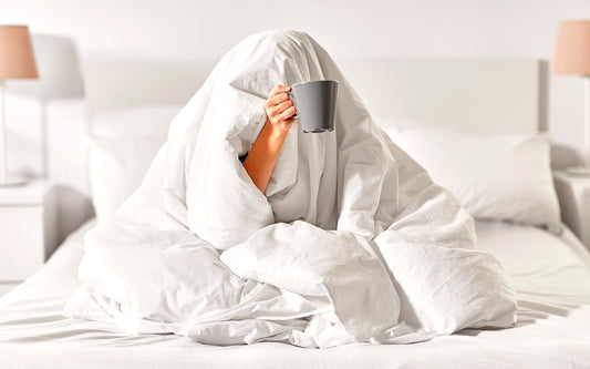 Frau unter einer weißen Bettdecke. Es guckt nur der Arm heraus, in dessen Hand eine Tasse ist. 