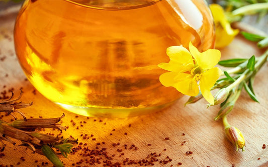 Ein Glasgefäß mit Öl gefüllt, Blüten der Nachtkerze und Pflanzensamen auf einem Holzbrett verstreut